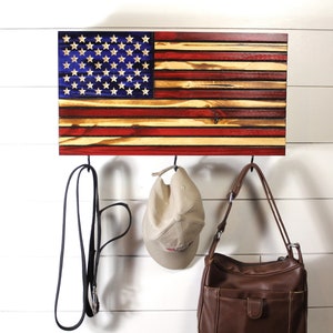 May include: Wooden American flag wall decor with three black metal hooks. The flag features red and natural wood stripes, a blue field with white stars. A black dog leash, a tan baseball cap, and a brown purse hang from the hooks.