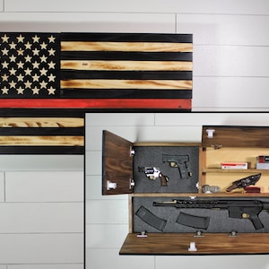 May include: Wooden American flag wall art with a red stripe, black stripes, and white stars. The flag opens to reveal a hidden compartment with firearms and ammunition. The cabinet is made of wood and has a natural finish.