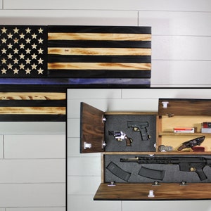 May include: A wooden American flag with a blue line, which is a symbol of support for law enforcement, is mounted on a wall. The flag is a secret compartment that opens to reveal a gun safe with a variety of firearms and ammunition.