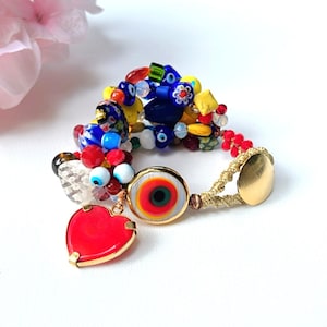 Puede incluir: Pulsera de cuentas multicolores con un dije de corazón rojo y un dije de ojo turco. La pulsera está hecha de cuentas de vidrio, incluyendo cuentas azules, rojas, amarillas y blancas. El dije de ojo turco es azul y blanco con un centro rojo.