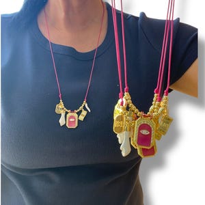 Puede incluir: Un collar de capas con múltiples dijes, incluyendo una pequeña casa, una mano y un rectángulo rosa con un ojo dorado. El collar está hecho de cuentas doradas y un cordón rosa.