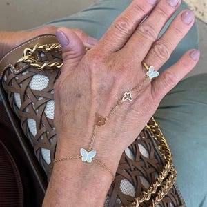 Puede incluir: Un conjunto de pulsera de cadena de mano y anillo dorados. La pulsera presenta una delicada cadena con dijes de mariposa y trébol. El anillo tiene un diseño de mariposa azul claro. La joya se usa con un bolso marrón y beige.