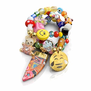 Puede incluir: Una colorida pulsera de cuentas con varios dijes. Los dijes incluyen un osito de peluche, una carita sonriente, un emoji de lágrimas y un colgante estampado. La pulsera presenta una mezcla de cuentas de vidrio en varias formas y colores.