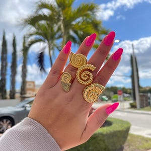 Puede incluir: Una mano muestra cuatro anillos dorados con diseños de conchas marinas. Los anillos varían en tamaño y forma, con intrincados patrones en espiral y texturizados. Las uñas están pintadas de color rosa brillante. El fondo incluye palmeras y un cielo nublado.