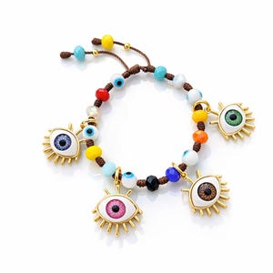 Puede incluir: Una pulsera de cuentas con abalorios dorados en forma de ojo. La pulsera tiene un cordón marrón e incluye cuentas azules, amarillas, rojas y blancas. Los abalorios de ojo tienen diferentes colores de iris, incluyendo azul, rosa y verde.