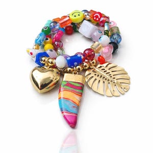 Puede incluir: Una colorida pulsera de cuentas con abalorios dorados. La pulsera presenta un corazón dorado, una hoja y un colgante a rayas. Las cuentas son de varios colores, incluyendo rojo, azul, amarillo y verde. La pulsera está sobre un fondo blanco.
