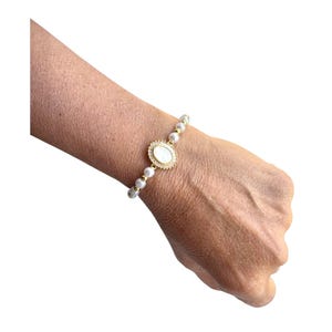 Puede incluir: Pulsera dorada con un colgante ovalado de piedra blanca, rodeado por un diseño de rayos de sol. La pulsera está ensartada con pequeñas perlas blancas redondas, creando un accesorio elegante y delicado.