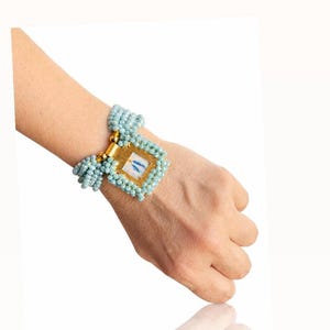 Puede incluir: Una pulsera de cuentas azul claro con una pieza central cuadrada dorada. La pieza central tiene un pequeño diseño rectangular con detalles en azul y blanco. La pulsera se lleva en la muñeca.