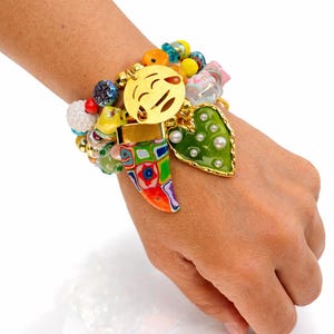 Puede incluir: Una colorida pulsera de cuentas con varios dijes, incluyendo un emoji dorado, un corazón verde con perlas y un dije en forma de cuerno multicolor. La pulsera presenta una mezcla de cuentas de varios colores y tamaños.