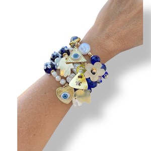Puede incluir: Una pulsera de múltiples hilos con varios dijes y cuentas. La pulsera incluye dijes dorados en forma de corazón, elefante y triángulo, cada uno con un diseño de ojo. Las cuentas son blancas, azules y color perla, con acentos en forma de flor.