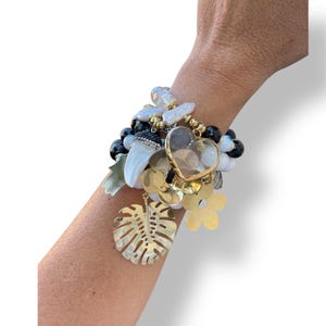 Puede incluir: Una pulsera de múltiples hilos con cuentas blancas, negras y doradas, junto con dijes dorados. Los dijes incluyen un corazón, flores y una hoja. La pulsera está diseñada para usarse en la muñeca.