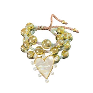 Puede incluir: Una pulsera en capas con orbes dorados y un colgante en forma de corazón. El colgante está delineado en dorado y adornado con pequeñas perlas blancas. La pulsera tiene una banda tejida multicolor y un cierre de lazo marrón ajustable.