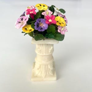 May include: A miniature arrangement of colorful pansies in shades of pink, yellow, purple, and white, atop a cream-colored, decorative column. The flowers are clustered together, creating a vibrant display. The column has a classical design.