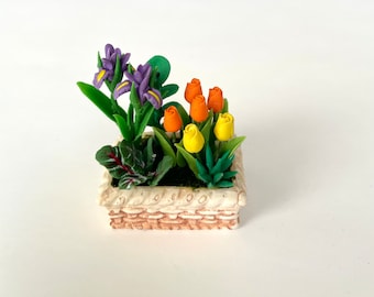 Puede incluir: Una jardinera decorativa en miniatura con un diseño tejido. La jardinera contiene iris moradas, tulipanes naranjas y amarillos y follaje verde. Las flores están dispuestas en una pequeña maceta cuadrada de color crema.