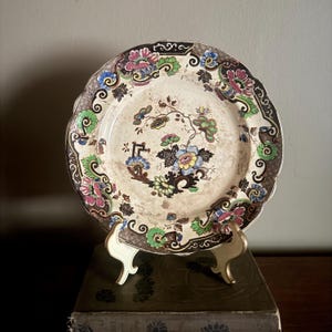May include: A round ceramic plate with a scalloped edge featuring a central floral motif in blue, yellow, and green. The border is decorated with dark brown, pink, and green floral patterns on a cream-colored background with visible aging.