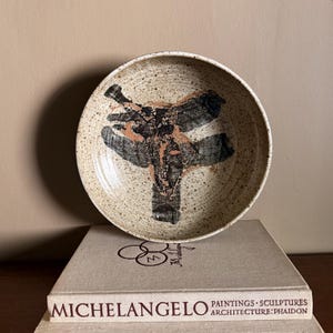 May include: A round ceramic bowl with a speckled cream-colored glaze featuring a central abstract design in black and copper tones. The bowl sits atop a beige book titled MICHELANGELO PAINTINGS SCULPTURES ARCHITECTURE PHAIDON.