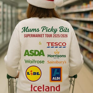 Op de afbeelding: Een witte trui met de tekst "Mums Picky Bits SUPERMARKET TOUR 2025/2026" en supermarktlogo's. De persoon draagt een rood-witte kerstmuts en duwt een winkelwagen.