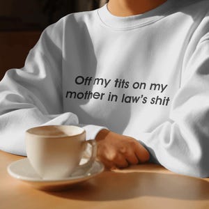 May include: White sweatshirt with the text "Off my tits on my mother in law's shit" in black. A white coffee cup and saucer are on a wooden table.
