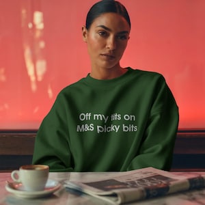 Könnte beinhalten: Ein dunkelgrünes Sweatshirt mit dem weißen Text "Off my tits on M&S picky bits". Das Sweatshirt wird von einer Person getragen. Eine Tasse Kaffee und eine Zeitung liegen auf dem Tisch vor der Person.