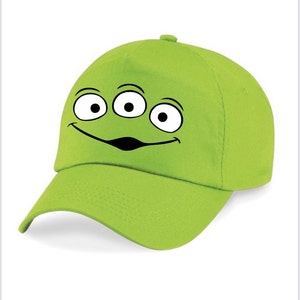 Puede incluir: Gorra de béisbol verde lima con la cara de un extraterrestre de dibujos animados. La cara tiene tres ojos y una boca sonriente, impresa en blanco y negro. La gorra tiene una visera curva y una corona redondeada.