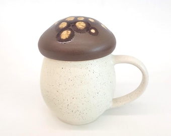 May include: A ceramic mug with a mushroom-shaped lid. The lid is dark brown with lighter brown spots. The mug is off-white with a speckled texture and a curved handle. The mug is designed to resemble a mushroom.
