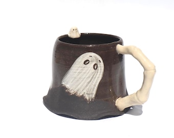 May include: A brown ceramic mug with a white ghost design and a bone-shaped handle. A small ghost figure sits on the rim. The mug has a dark brown exterior with a lighter brown interior. The ghost design is white with black eyes.