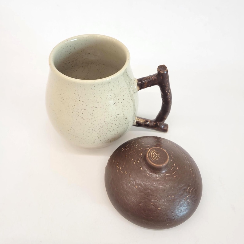 May include: A ceramic mug with a speckled, off-white finish and a dark brown, branch-shaped handle. The mug is accompanied by a matching lid. The lid is a dark brown color with a textured surface and a small, circular knob.