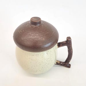 May include: A ceramic mug with a brown acorn-shaped lid and a twig-like handle. The mug is off-white with speckled detailing. The lid has a textured surface and a small, circular knob.