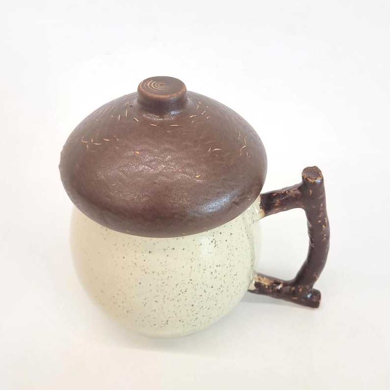 May include: A ceramic mug with a brown acorn-shaped lid and a twig-like handle. The mug is off-white with speckled detailing. The lid has a textured surface and a small, circular knob.