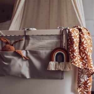 May include: A white crib with a gray and white fabric organizer with a brown stuffed animal inside. A rainbow macrame wall hanging is attached to the crib. A brown and white patterned muslin swaddle is hanging on the side of the crib.