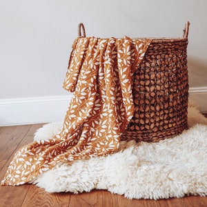 May include: A brown wicker basket with a white and mustard yellow patterned muslin blanket draped over the top.