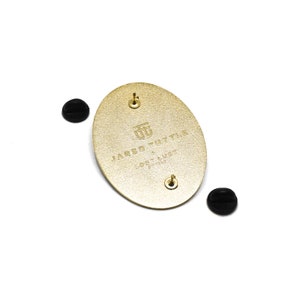 May include: Gold oval-shaped pin with the text "JARED TUTTLE LOST + FOUND" and a logo of a stylized "JT" in the center.