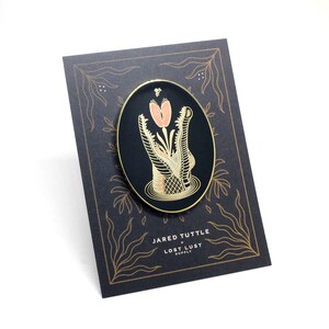 May include: Gold enamel pin featuring a black and white alligator with a pink flower in its mouth. The pin is on a black background with gold floral designs. The text "JARED TUTTLE + LOST LUST SUPPLY" is printed below the pin.