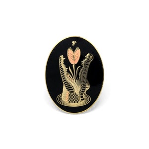 May include: A black and gold enamel pin featuring a stylized alligator with its mouth open, a pink flower growing from its mouth, and a small bee flying above the flower.