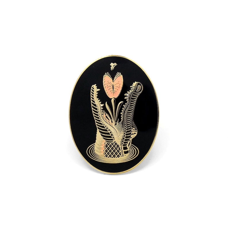 May include: A black and gold enamel pin featuring a stylized alligator with its mouth open, a pink flower growing from its mouth, and a small bee flying above the flower.