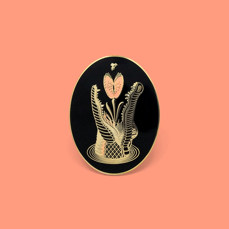 May include: Black and gold enamel pin featuring a crocodile with its mouth open, swallowing a pink flower. The pin has a black background and gold accents.