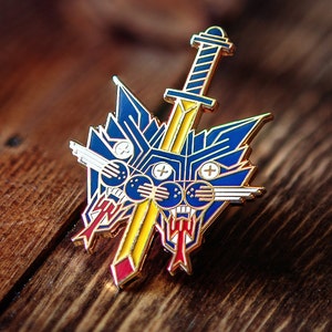 May include: A detailed enamel pin featuring a stylized sword overlaid on a blue and gold emblem. The pin has a yellow sword with a blue handle, and the emblem has red and white accents. The pin is set against a wooden background.