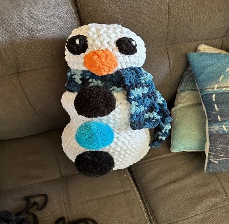 Pode incluir: Uma almofada de boneco de neve de croch&ecirc; branco com olhos pretos, nariz laranja e uma echarpe azul e branca. O boneco de neve est&aacute; sentado em um sof&aacute; marrom.
