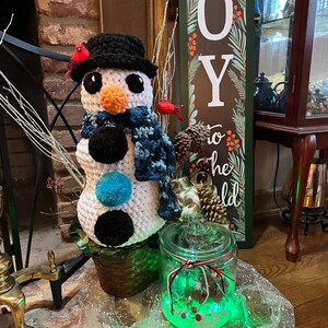 Pode incluir: Um boneco de neve de croch&ecirc; com um chap&eacute;u preto e uma echarpe azul est&aacute; em uma cesta de vime ao lado de um frasco de vidro com uma luz verde dentro. O frasco &eacute; decorado com uma fita vermelha e branca. O fundo apresenta uma lareira de tijolos e uma placa de madeira que diz "JOY to the world".