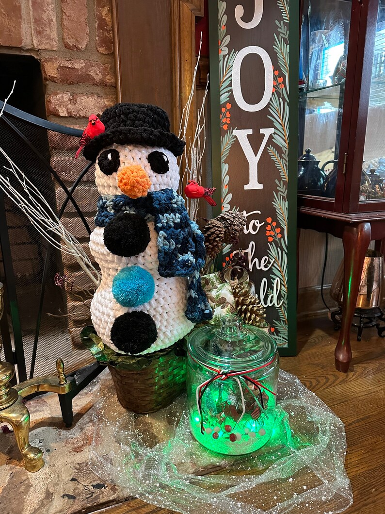 Pode incluir: Um boneco de neve de croch&ecirc; com um chap&eacute;u preto e uma echarpe azul est&aacute; em uma cesta de vime ao lado de um frasco de vidro com uma luz verde dentro. O frasco &eacute; decorado com uma fita vermelha e branca. O fundo apresenta uma lareira de tijolos e uma placa de madeira que diz "JOY to the world".