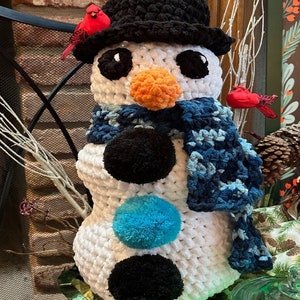 Pode incluir: Um boneco de neve de croch&ecirc; branco com um chap&eacute;u preto, um cachecol azul e pompons pretos e azuis. O boneco de neve tem um nariz de cenoura laranja e olhos pretos.