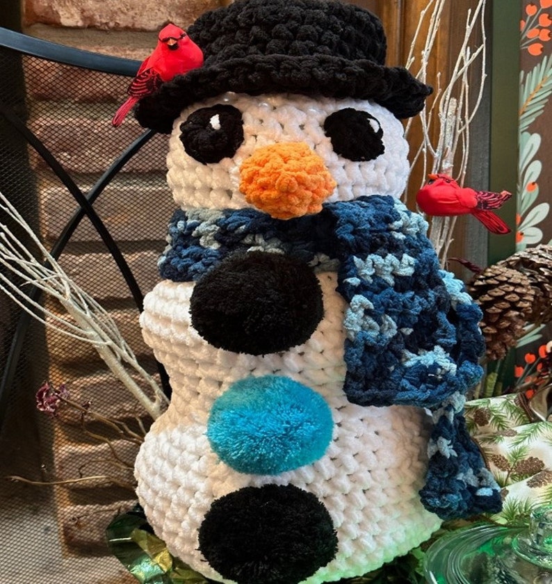 Pode incluir: Um boneco de neve de croch&ecirc; branco com um chap&eacute;u preto, um cachecol azul e pompons pretos e azuis. O boneco de neve tem um nariz de cenoura laranja e olhos pretos.