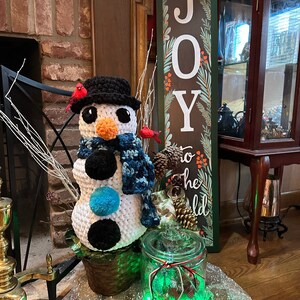 Pode incluir: Um boneco de neve de croch&ecirc; feito com fio branco e preto, com uma echarpe azul e bot&otilde;es pretos. O boneco de neve est&aacute; sentado em uma cesta de vime marrom com cardeais vermelhos empoleirados na borda. Um frasco de vidro com luzes verdes e neve branca est&aacute; em primeiro plano. Uma placa de madeira com a palavra "ALEGRIA" est&aacute; no fundo.