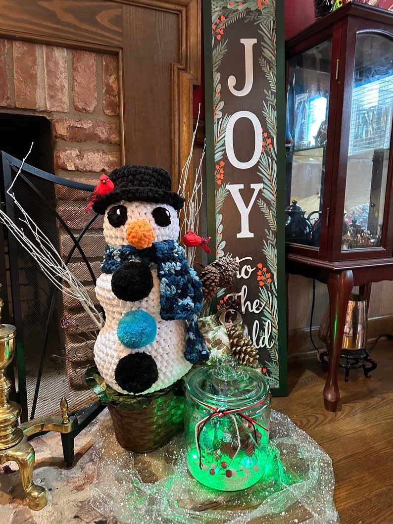 Pode incluir: Um boneco de neve de croch&ecirc; feito com fio branco e preto, com uma echarpe azul e bot&otilde;es pretos. O boneco de neve est&aacute; sentado em uma cesta de vime marrom com cardeais vermelhos empoleirados na borda. Um frasco de vidro com luzes verdes e neve branca est&aacute; em primeiro plano. Uma placa de madeira com a palavra "ALEGRIA" est&aacute; no fundo.