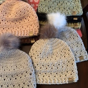 Può includere: Sei berretti all'uncinetto con pon pon in vari colori, tra cui beige, grigio, rosa e verde. Ogni berretto ha un piccolo cartellino dorato.