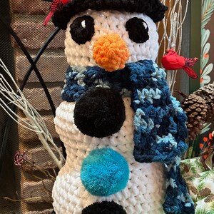 Pode incluir: Um boneco de neve de croch&ecirc; branco com um chap&eacute;u preto, uma echarpe azul e pompons pretos e azuis. Dois p&aacute;ssaros cardeais vermelhos est&atilde;o empoleirados no chap&eacute;u do boneco de neve.