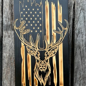 May include: Wooden wall art featuring an American flag design with a deer silhouette. The flag has a black background with light brown stripes and stars. The deer is intricately carved, showcasing the antlers and facial features.