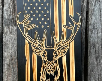 May include: Wooden wall art featuring an American flag design with a deer silhouette. The flag has a black background with light brown stripes and stars. The deer is intricately carved, showcasing the antlers and facial features.