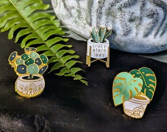 May include: Three enamel pins featuring potted plants. The first pin is a green and white succulent in a white pot with gold accents. The second pin is a green plant in a white pot on a gold stand with the text "crazy plant lady". The third pin is a green monstera plant in a white pot with gold accents.