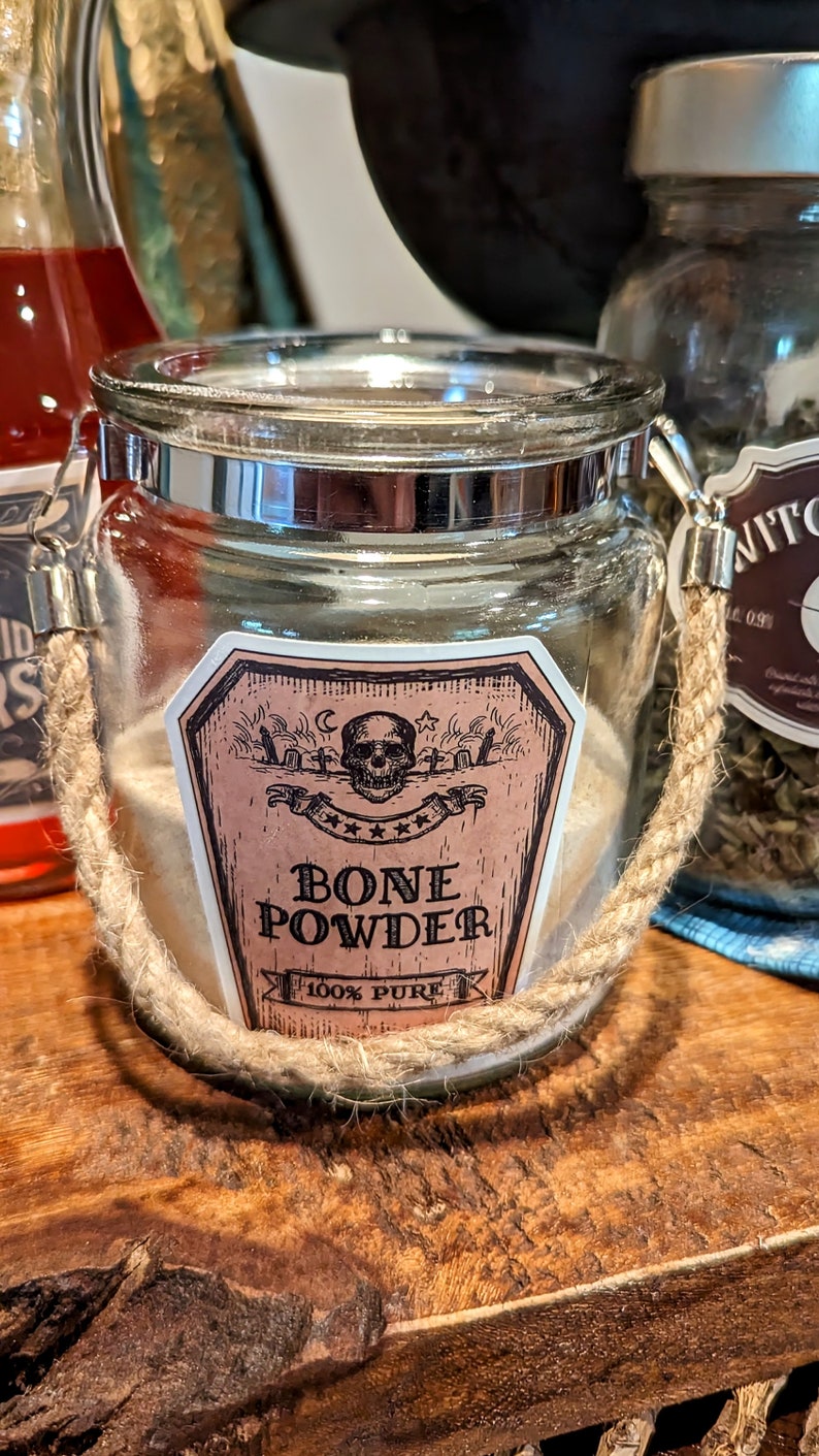 May include: A glass jar with a rope handle filled with a white powder. The jar has a label that says "Bone Powder 100% Pure" with a skull and crossbones graphic.